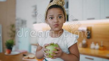 漂亮的非裔美国小女孩，卷发，手里拿着绿苹果，坐在桌子上，阳光明媚